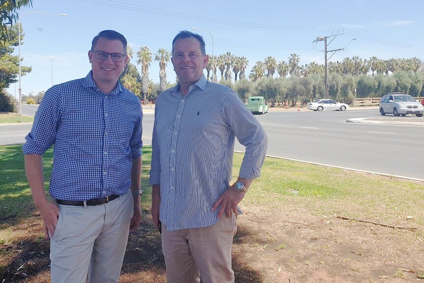 Two men stand smiling at the camera. There is a road intersection with cars driving behind them.