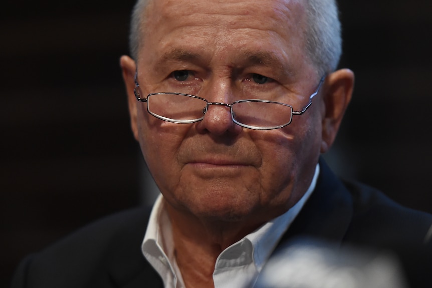 A close-up of a balding man in his 70s wearing frameless glasses and a suit, who has a sombre expression. 