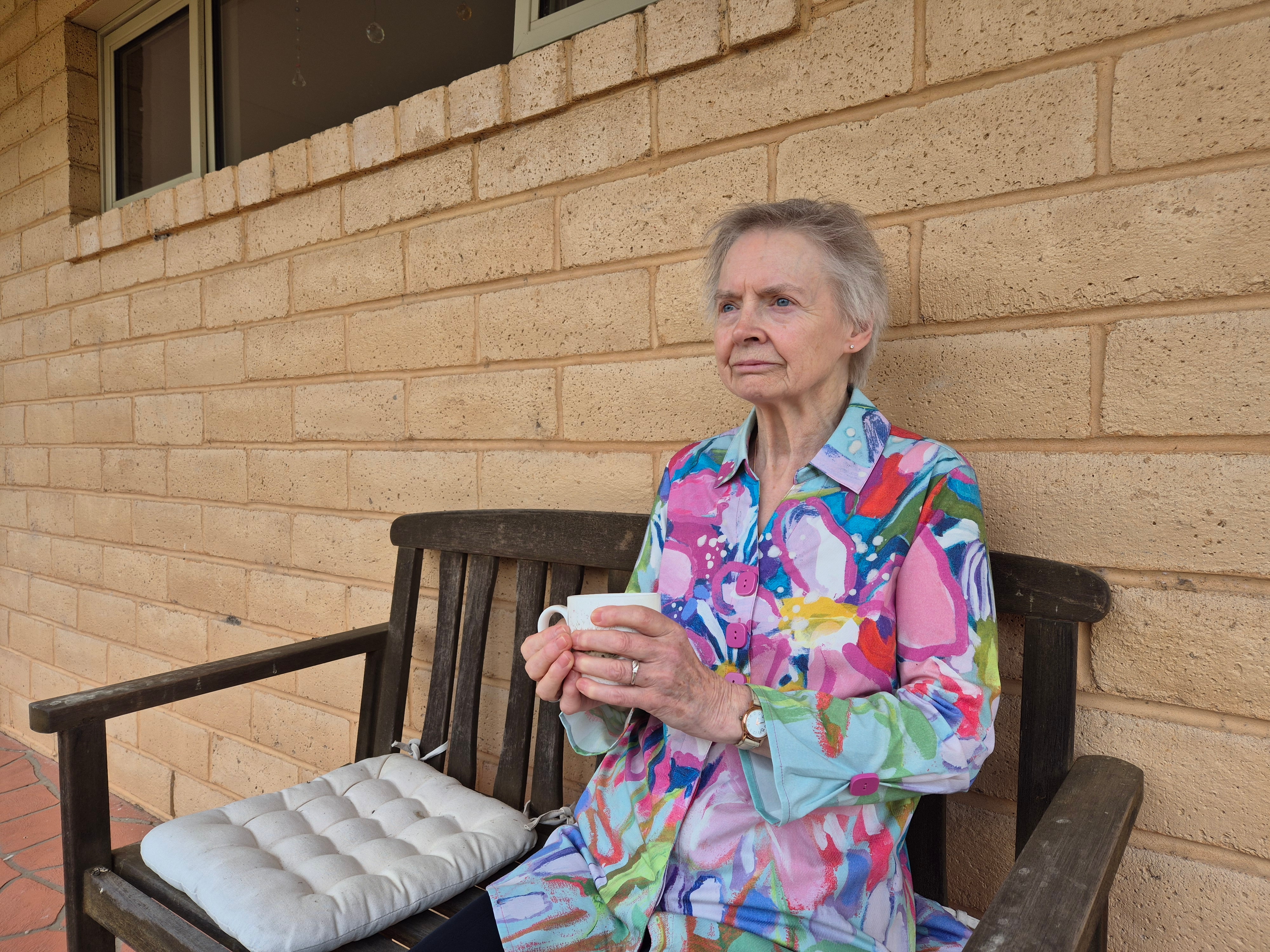 Uma senhora idosa sentada do lado de fora de uma casa de tijolos amarelos segurando uma xícara de chá.