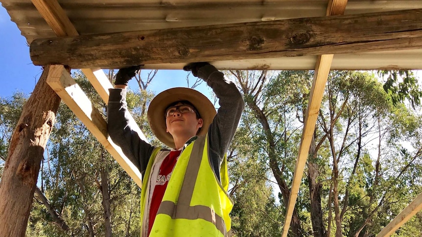 A boy in high-visibility workwear holds up a roof during construction.