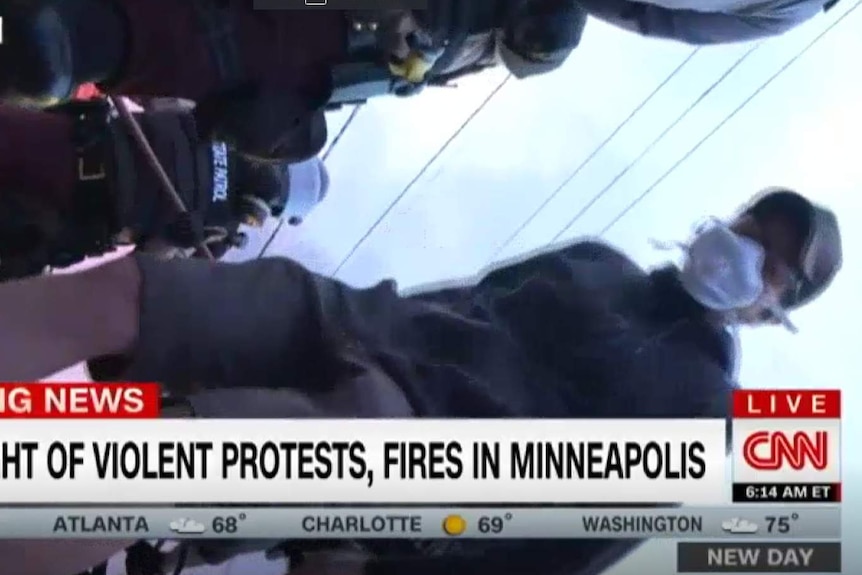 A sideways frame taken from a live news feed shows a person in a face mask and cap looking into the camera.