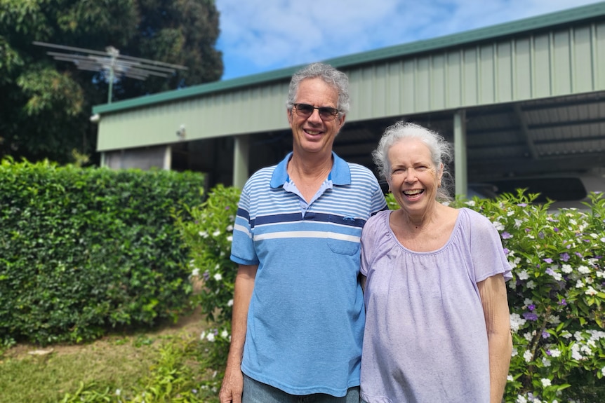 A couple in their sixties stand in front of a green hedge with their house in the background.