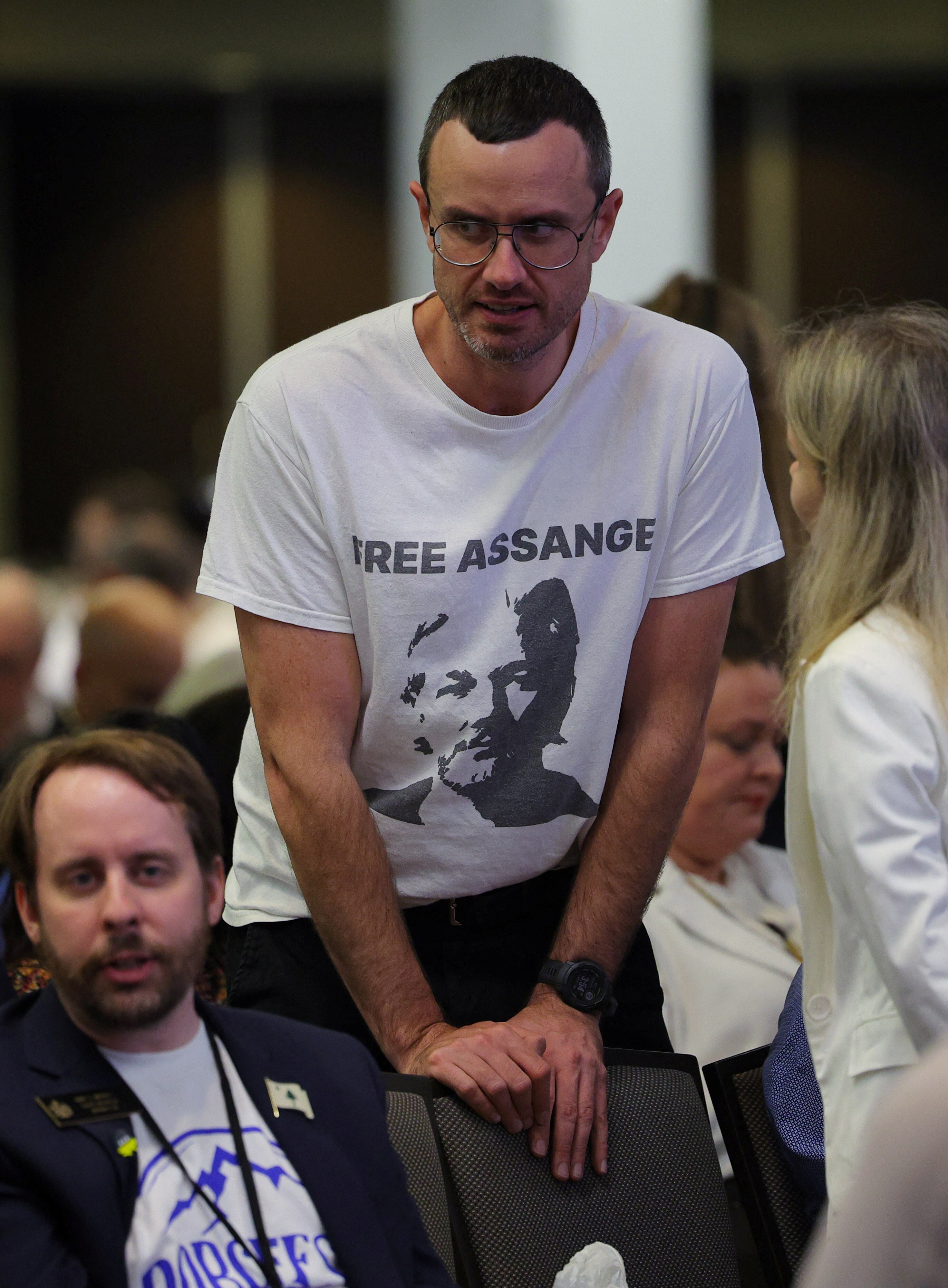 Gabriel Shipton speaks to someone at a conference wearing a shirt with his brother's face on it.