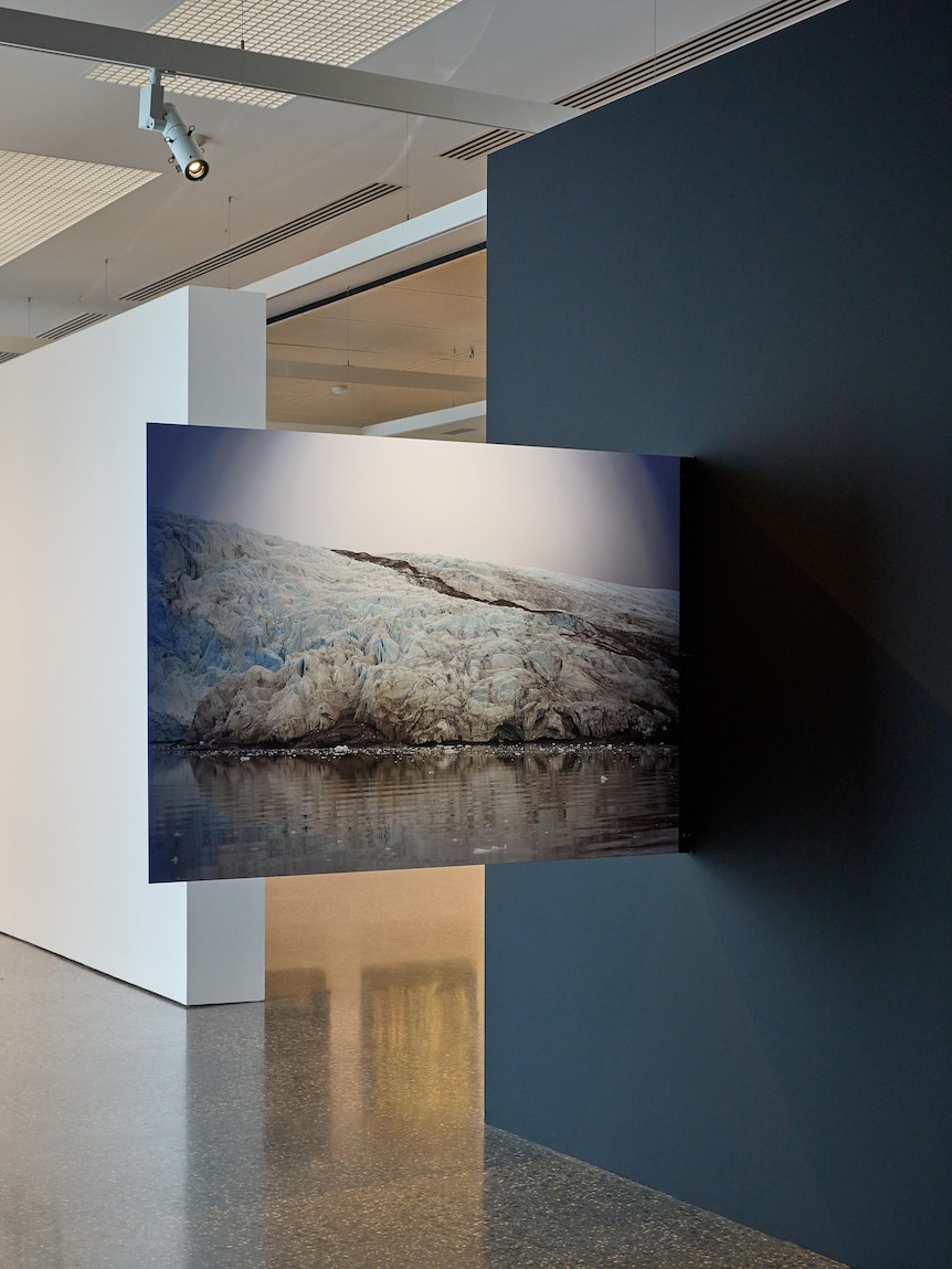 In a gallery, a photograph of a melting glacier juts out from the wall at a 90-degree angle.