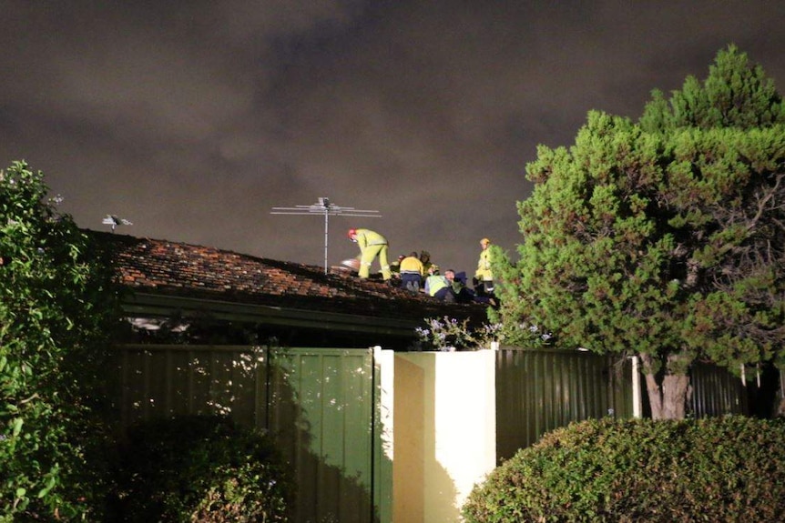 Emergency services workers in fluoro jackets on the tiled roof of a Yokine house at night.