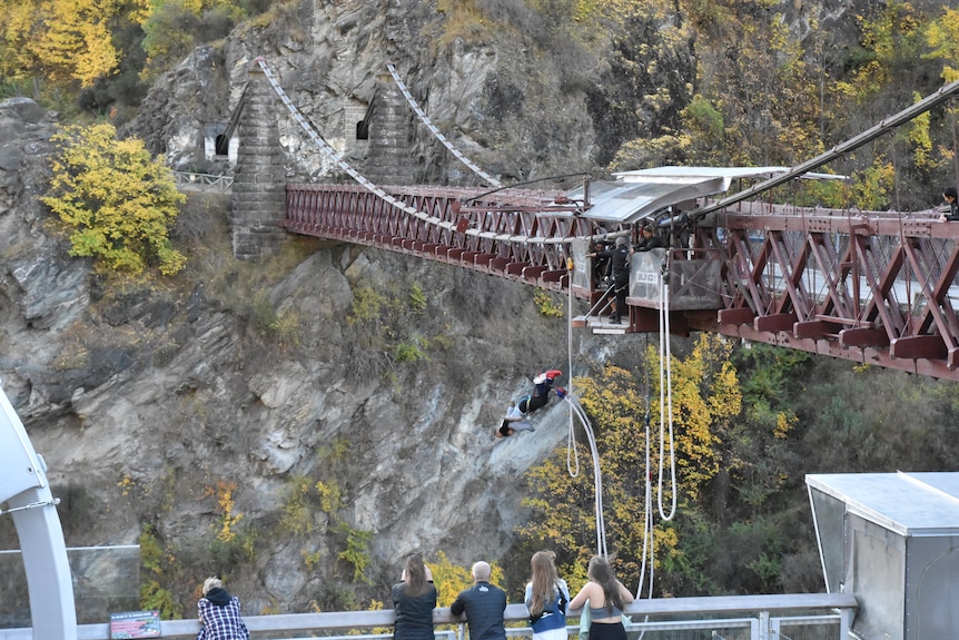 A woman and a man hold each other after bungee-jumping from a suspension bridge with five people watching their fall.
