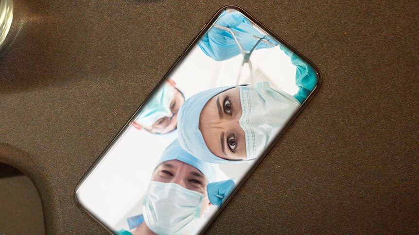 A phone sits on a desk - inside the phone three surgeons in marks look out as if they are operating.