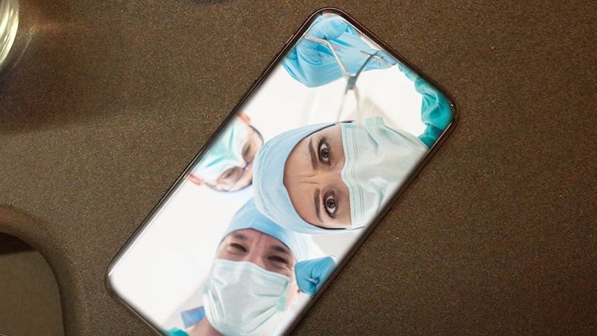 A phone sits on a desk - inside the phone three surgeons in marks look out as if they are operating.