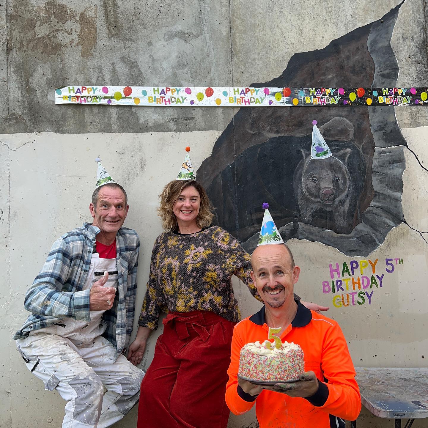 Three people wearing party hats stand in front of a mural of a wombat, one of them holds a cake.