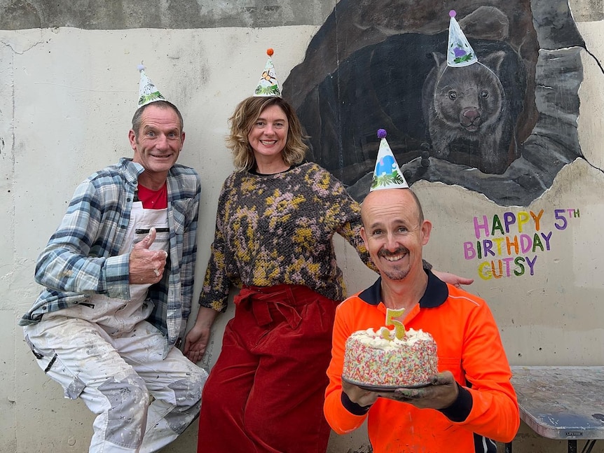 Three people wearing party hats stand in front of a mural of a wombat, one of them holds a cake.