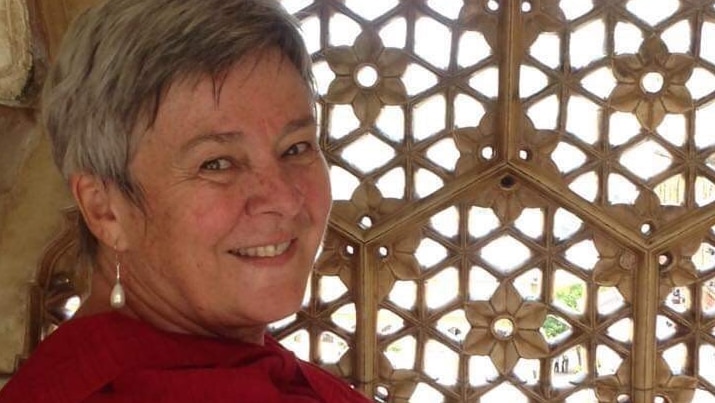 A woman in black print shirt and red scarf holds a decorative wooden wall while turning to face camera, smiling.