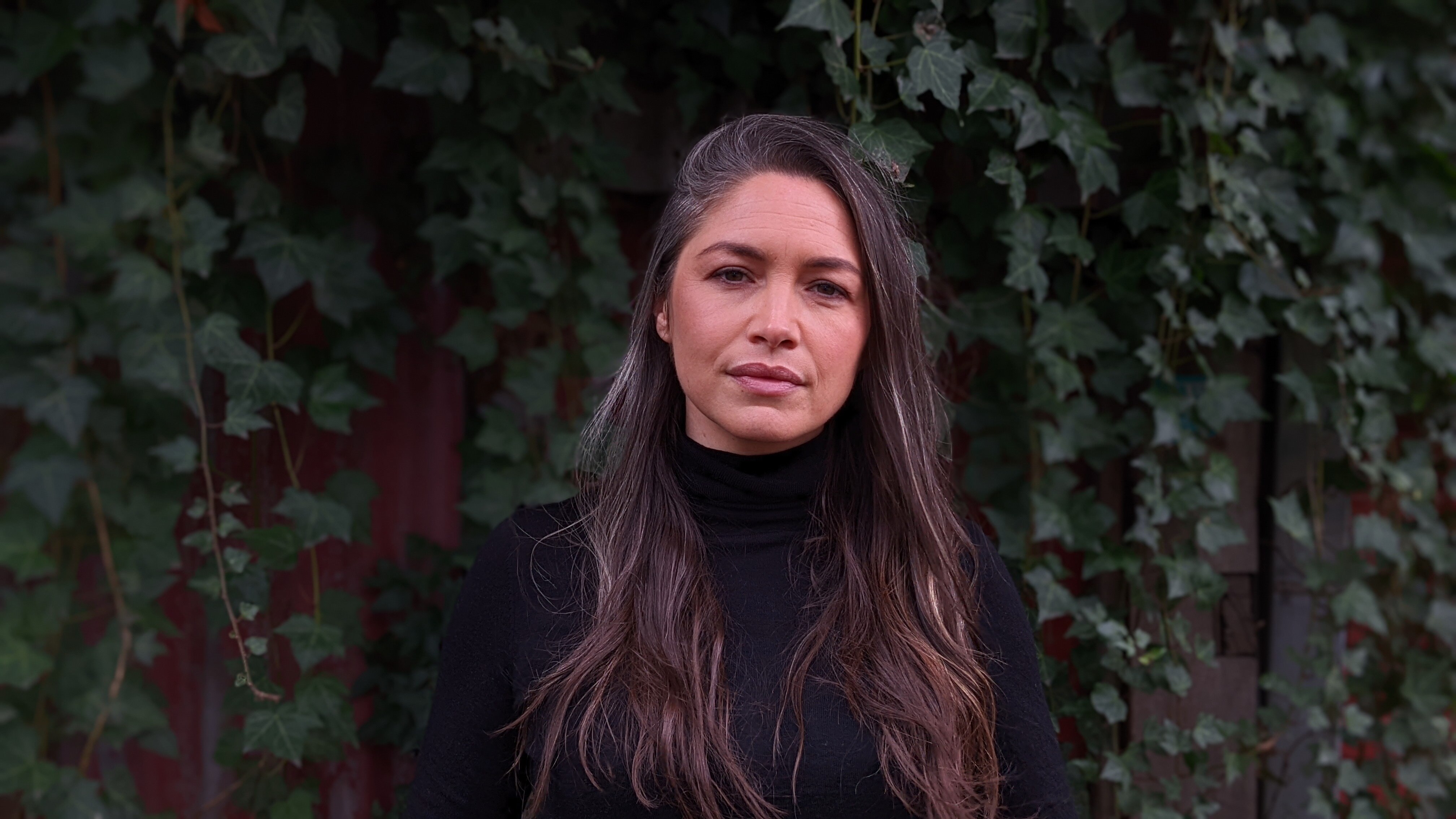 A woman wearing black with long brown hair stands in front of a background with green hanging plants. 