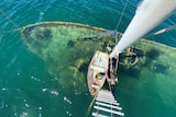 Birdseye view of sunken boat undewater, taken from crows nest high above, with ladder, mast, and small tinny below