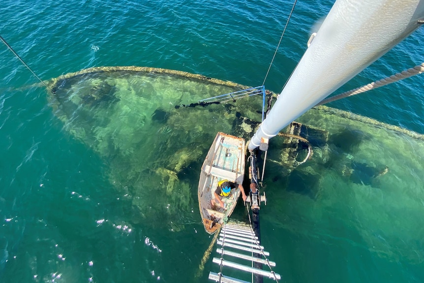 Birdseye view of sunken boat undewater, taken from crows nest high above, with ladder, mast, and small tinny below