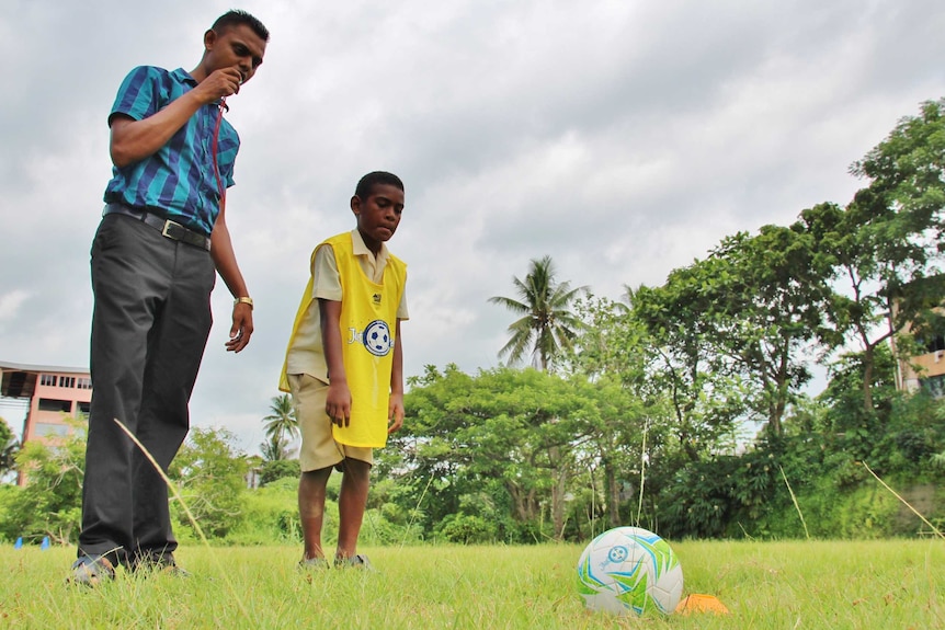 A male teacher stands next to a boy in a yellow bib who is preparing to run and kick the ball.