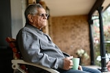 Older man with grey hair and glasses sits on his porch with a cup of tea in hand