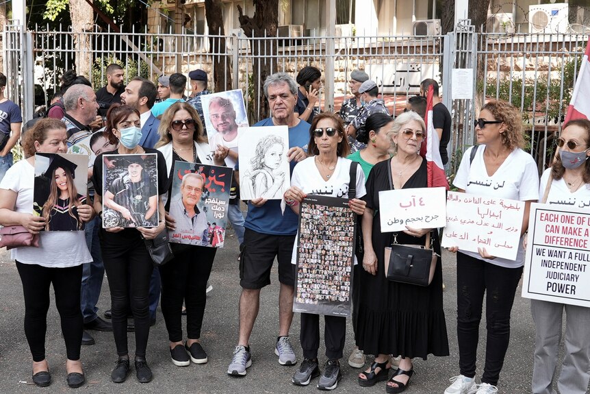 A group of mostly middle-aged people hold photos and drawings of lost loved ones in front of a gated building.