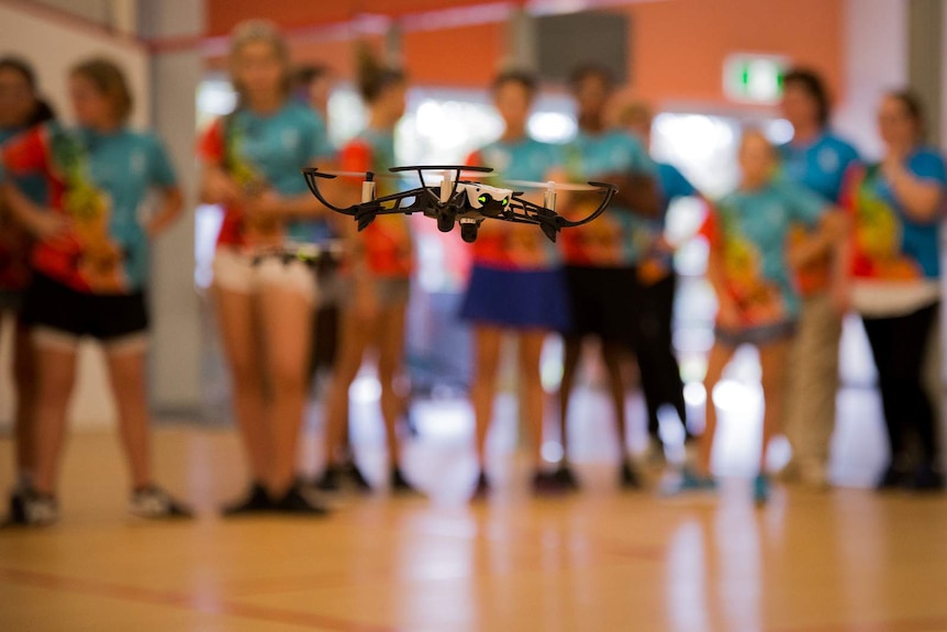 A small drone hovers in mid air as a crowd of girls aged between 10 and 17 look on in the background.