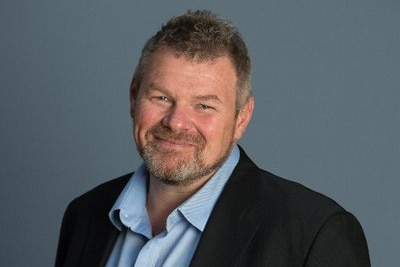 A profile shot of a smiling man wearing a blue shirt and a black blazer.