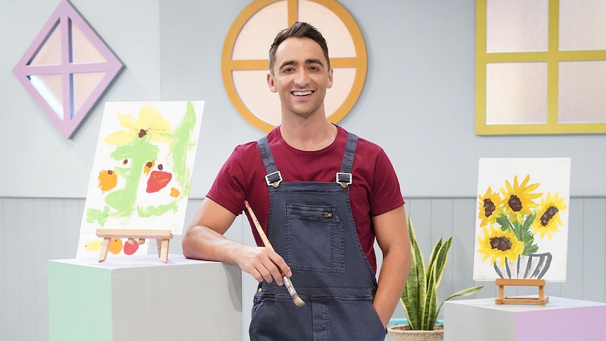 Matt stands holding a paintbrush in front of paintings of sunflowers