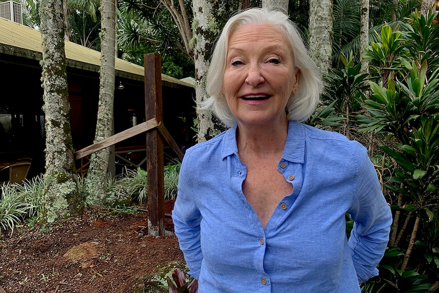 An older woman in a blue shirt standing outside on a treed property, smiling.