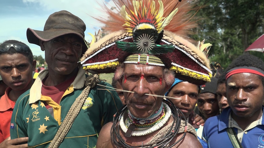 A man wearing traditional Huli wigman clothing and headdress, with long thin stakes through his nose, stares into the camera