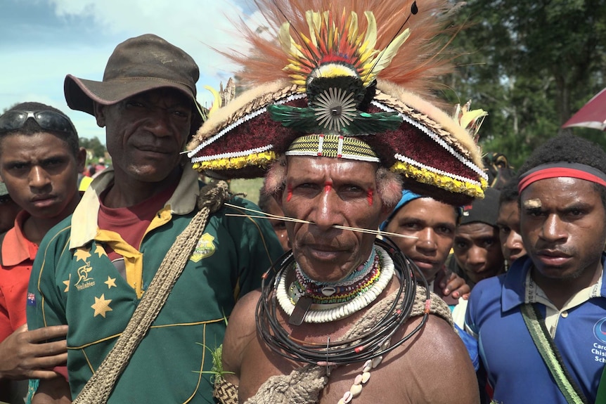 A man wearing traditional Huli wigman clothing and headdress, with long thin stakes through his nose, stares into the camera