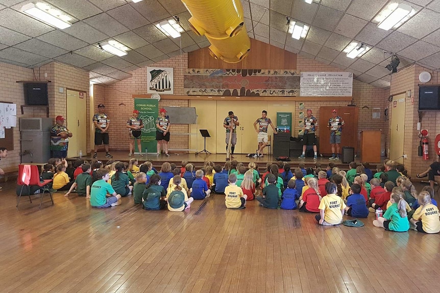 A man plays a didgeridoo on stage for children sitting on the floor of a school hall.