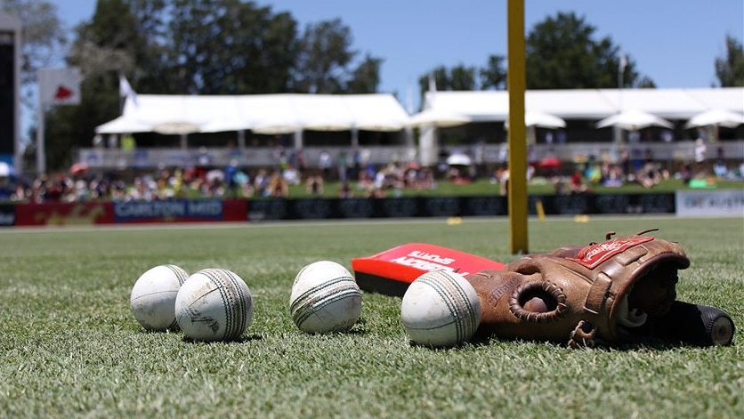 Cricket equipment including balls, a glove and bat lying on the ground of an oval.