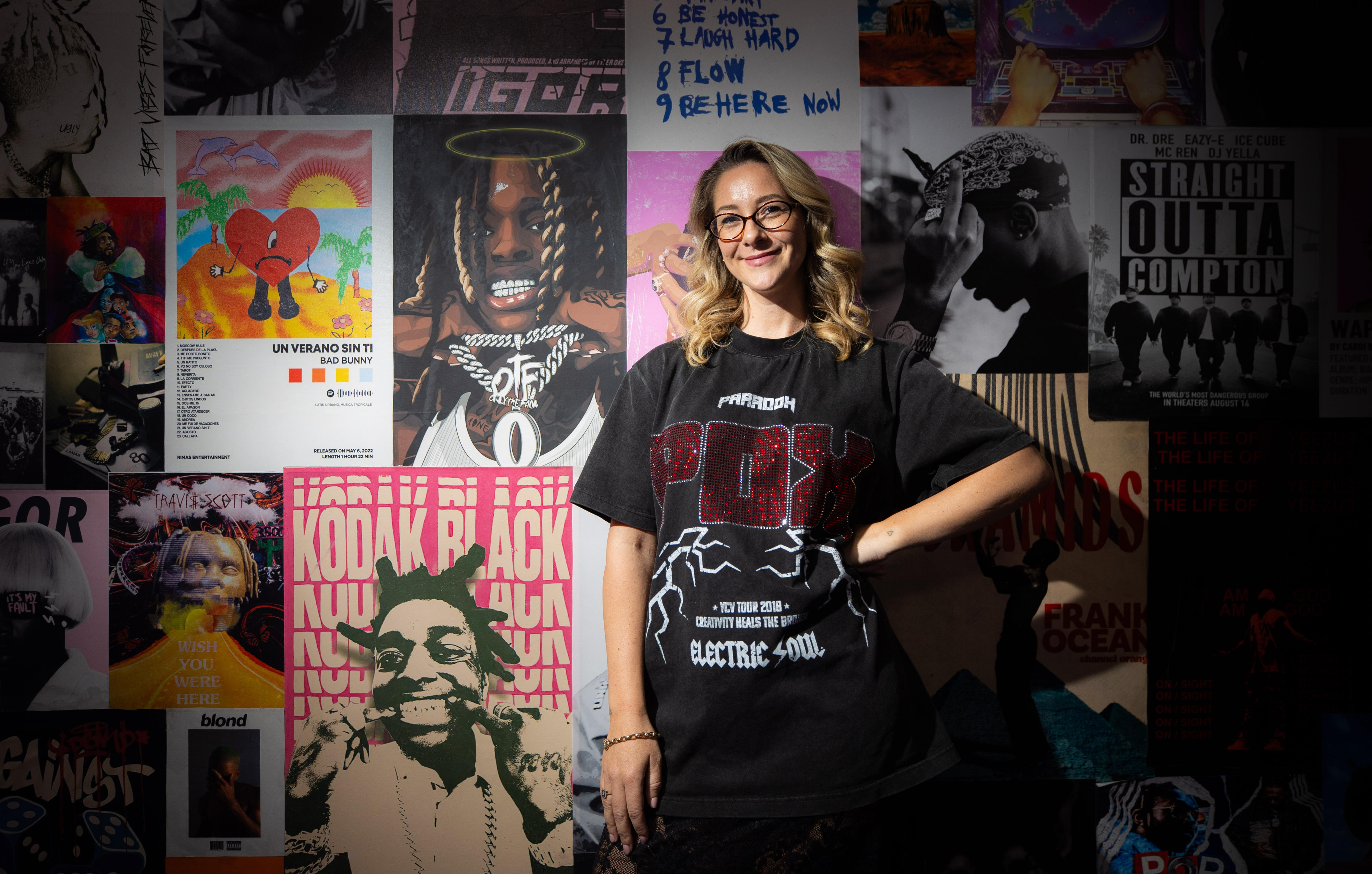 Una mujer con una camiseta negra se para frente a una pared con carteles musicales