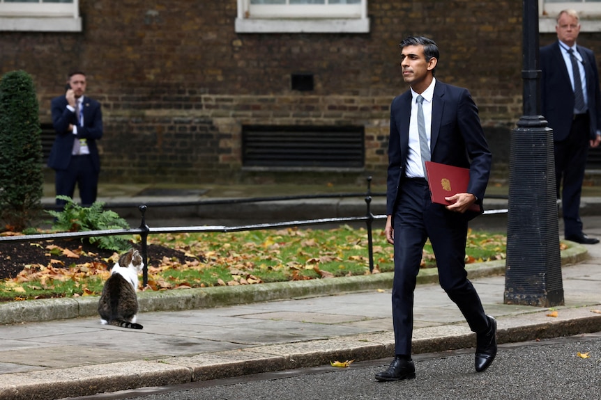 rishi sunak walks down the road holding a red folder. larry the cat is sitting on the footpath looking away from him