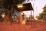 A man sits outside at dusk talking on a public telephone