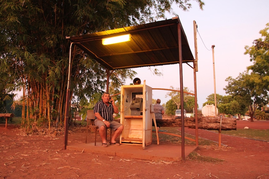 A man sits outside at dusk talking on a public telephone