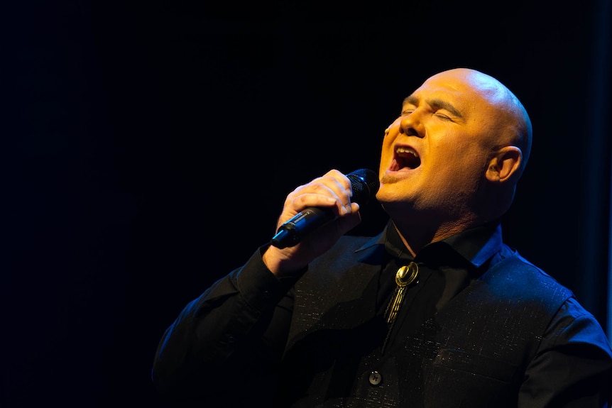 A man singing expressively under warm stage lights.