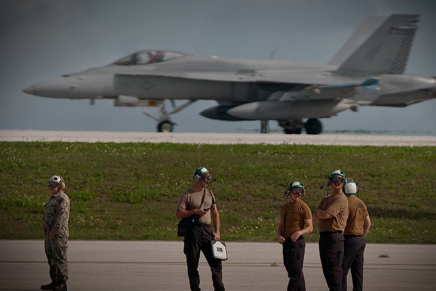 Personnel in khaki Tshirts and camouflage pants stand on a tarmac. Behind them is a fighter jet