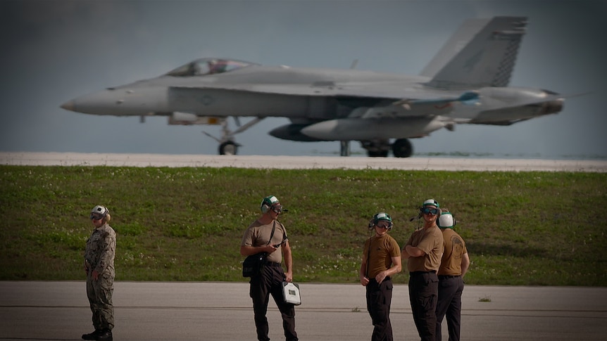 Personnel in khaki Tshirts and camouflage pants stand on a tarmac. Behind them is a fighter jet