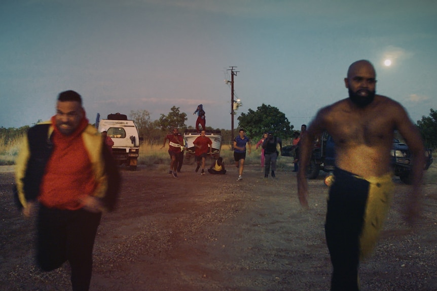 Two Aboriginal men running in the foreground and a group of others running in the background at twilight