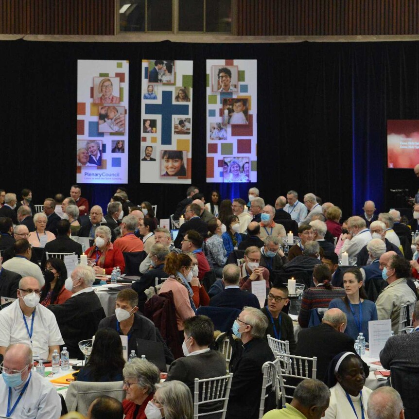 crowd sits around different tables in hall, in front is a stage with long posters of people's faces, and a cross in the middle