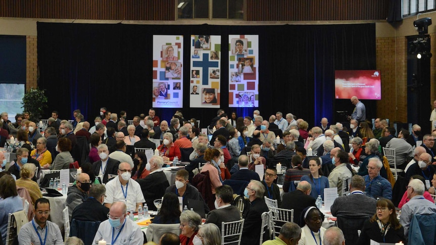 crowd sits around different tables in hall, in front is a stage with long posters of people's faces, and a cross in the middle