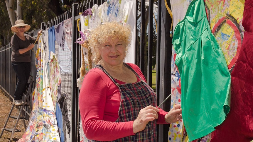 Artist holding a small paintbrush stands in front of an installation of fabric and clothing hung on a black school fence