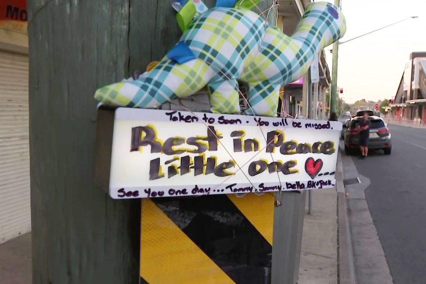 A dinosaur toy sits with a sign that reads "Rest in peace little one" are adhered to a pole by the roadside.