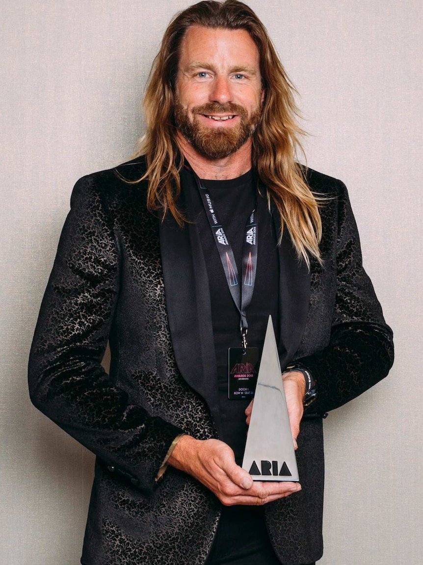 Scott Maxwell standing against a grey background holding his ARIA award. He is wearing a suit and smiling.