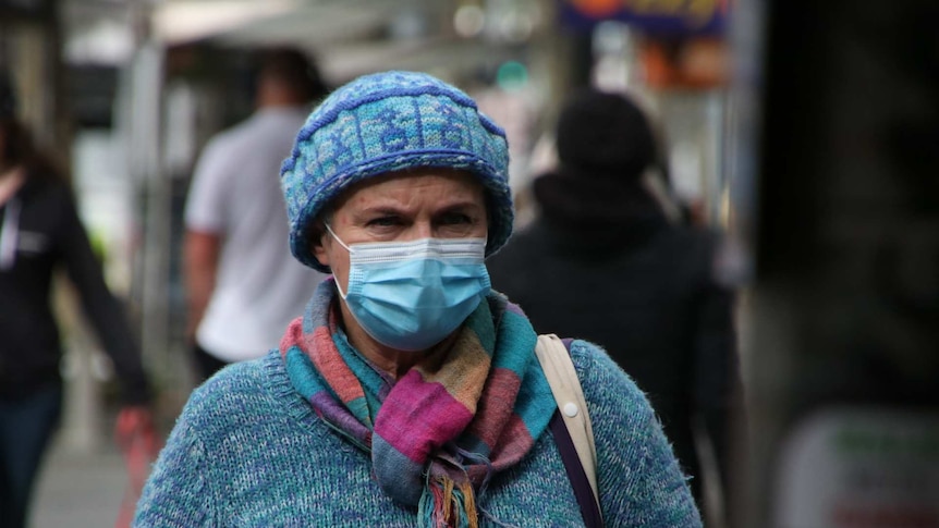 A close-up of a woman wearing a blue surgical mask, beanie and woollen jumper
