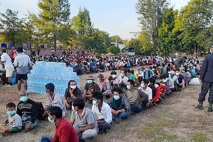 People sit in long rows on the ground next to a stack of water bottles. 