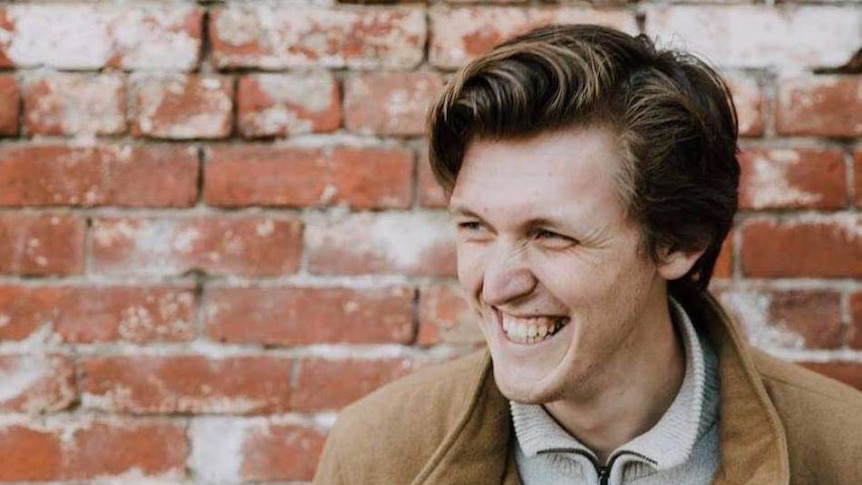 A young man poses for a photo in front of a brick wall, he is smiling and reflecting on work life balance.