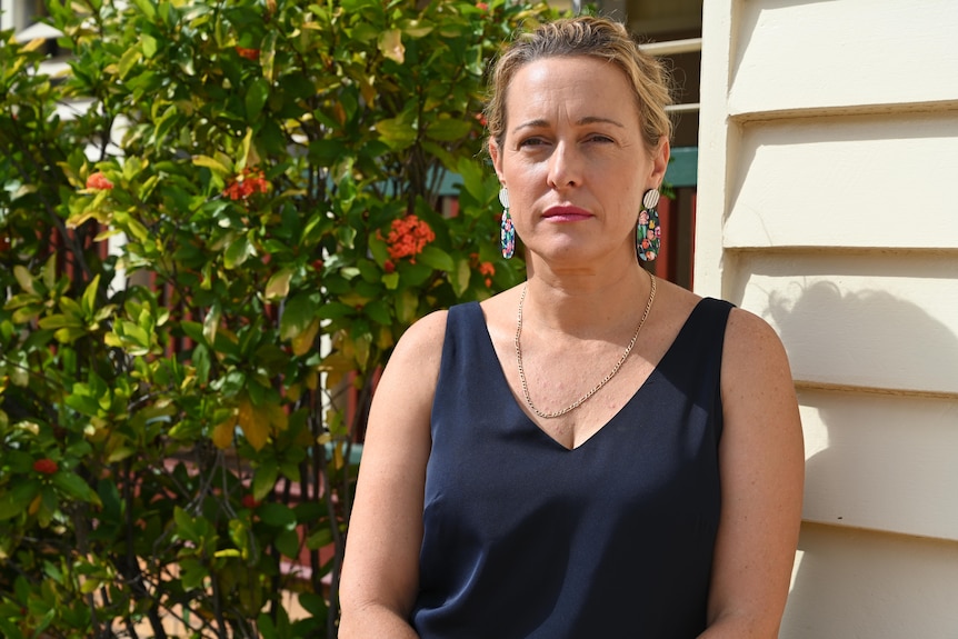 A woman in a sleeveless blouse stands beside a building in Gayndah next to a red flowering tree.