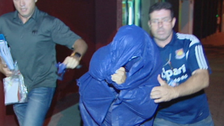 A man wearing a blue jumpsuit with his face covered is flanked by two plain clothes police officers as they escort him.