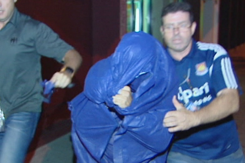 A man wearing a blue jumpsuit with his face covered is flanked by two plain clothes police officers as they escort him.