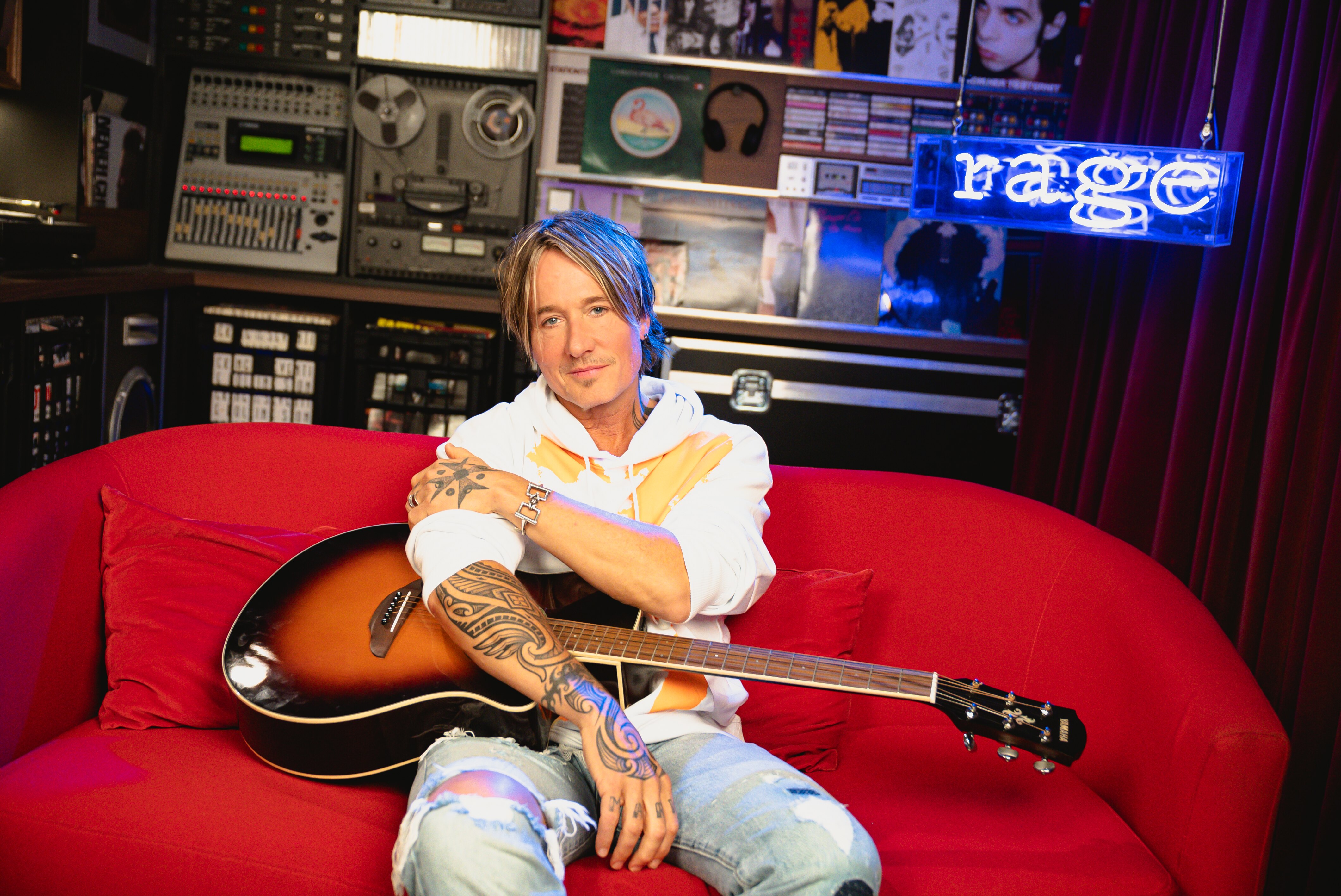Keith urban sits on a red couch with a guitar lying across his lap