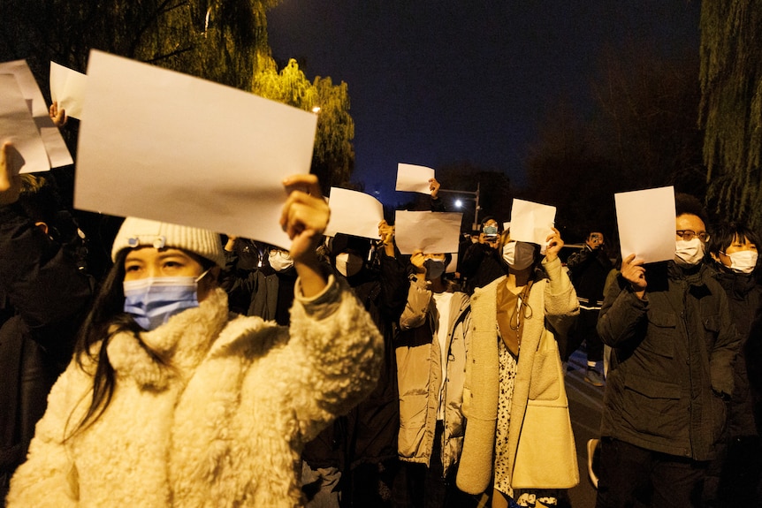 People hold white sheets of paper as they gather in a street in protest.